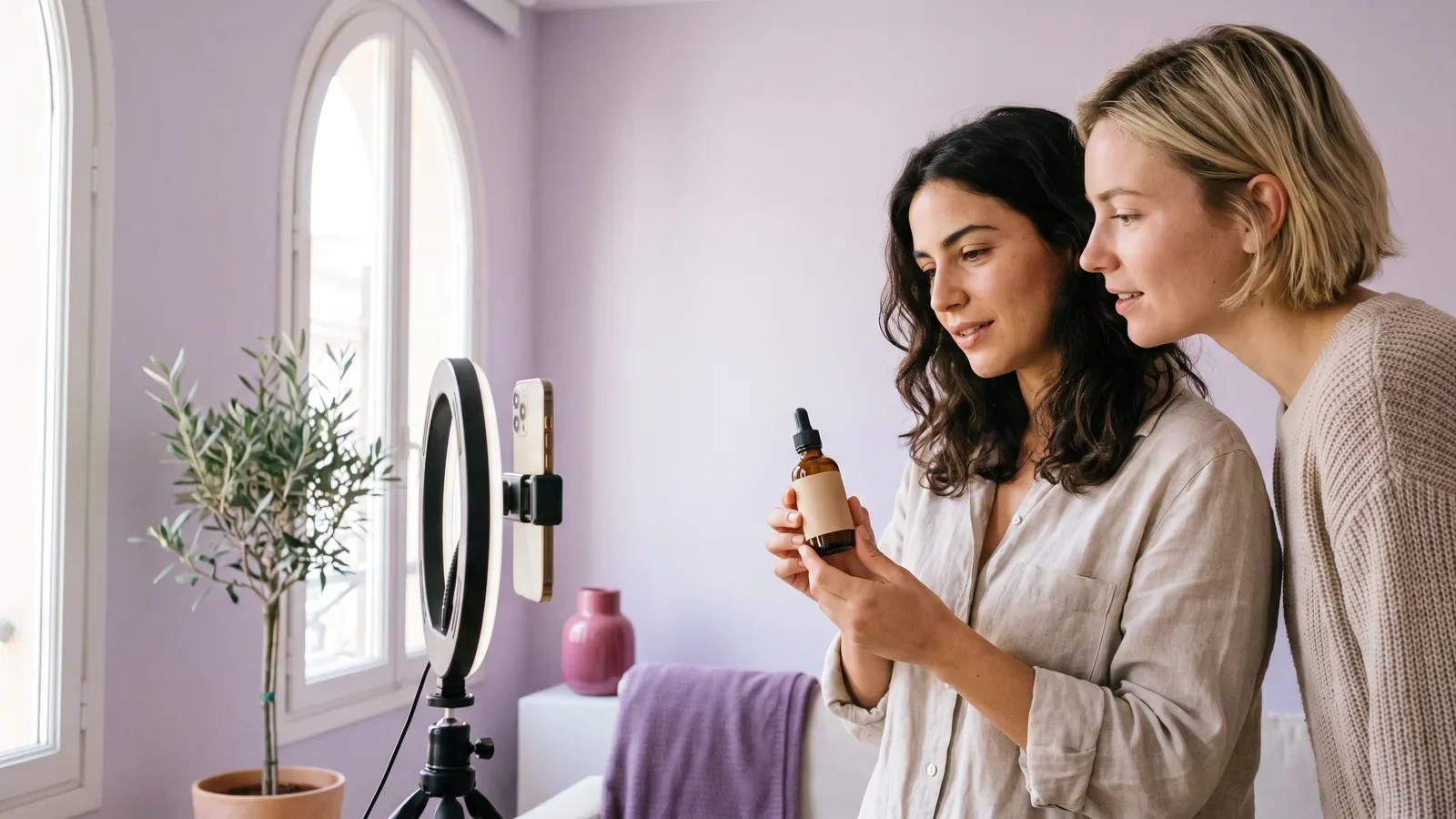 Two women filming a vertical video of a skincare serum with a smartphone on a ring light in a bright lilac room
